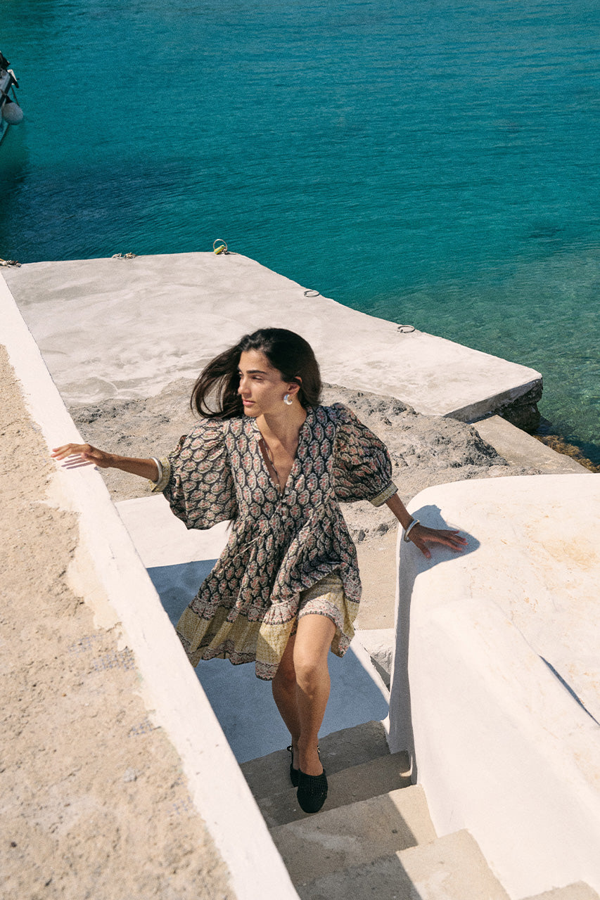 Woman on a seaside stone ledge overlooking turquoise water wearing the Daughters of India Bhoomi Mini Dress in Onyx, the dark block print fabric contrasting with the bright sea