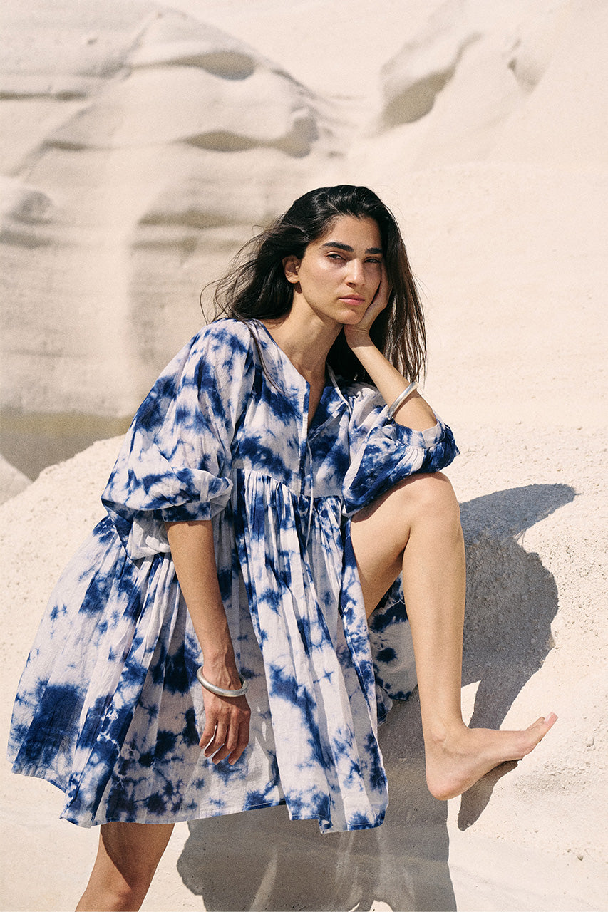 Kyra Mini Dress Deep Sea — seated on white sand dune, deep sea shibori tie-dye cotton mini dress with V-neckline and balloon sleeves, coastal beach setting | Daughters of India