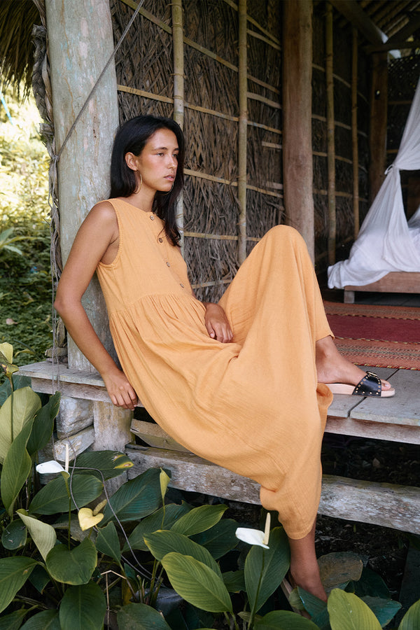 Nadi Dress Amber — seated on tropical hut step, amber double gauze cotton midi dress showing flowing skirt drape, thatched walls and greenery | Daughters of India