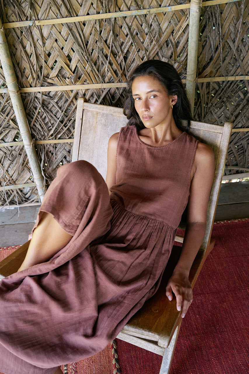 Nadi Dress Chocolate — seated in wooden chair in bamboo hut, chocolate brown double gauze cotton midi dress draped casually, woven palm wall and red rug | Daughters of India