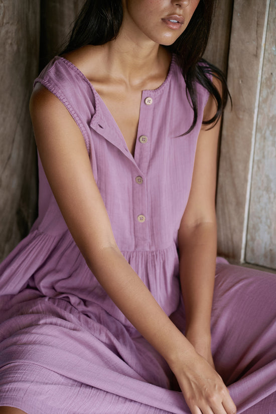 Nadi Dress Mauve — seated close-up showing wooden button front and gathered waist, mauve double gauze cotton midi dress draped over legs, rustic wooden wall background | Daughters of India