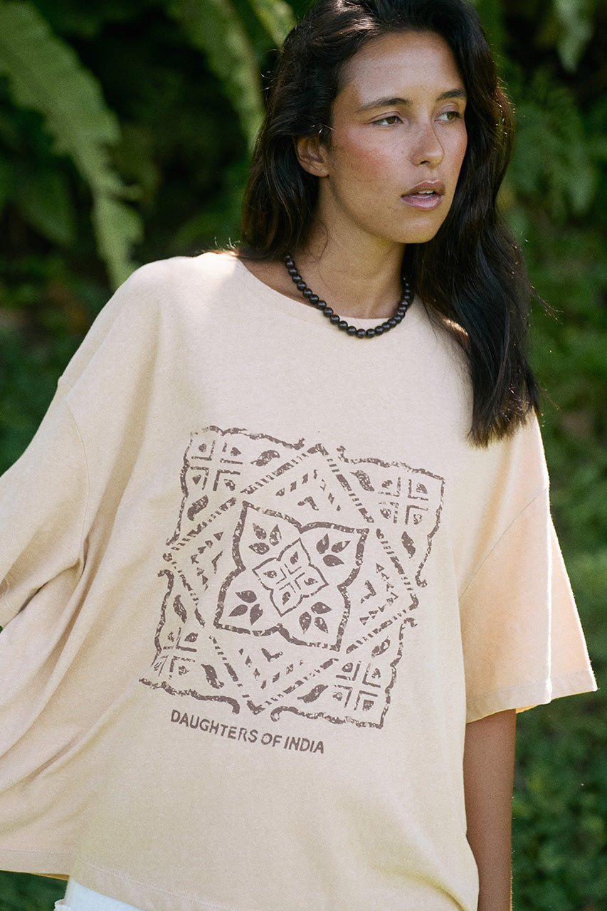 Woman wearing the Daughters of India Shanti Tee in Cashew outdoors against lush green foliage, featuring the block print design and brand text